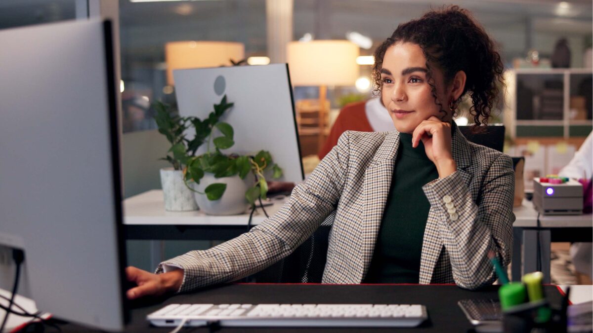 Image shows a marketer preparing for Black Friday. A young woman wearing a blazer looks confidently at her computer, in an office environment.